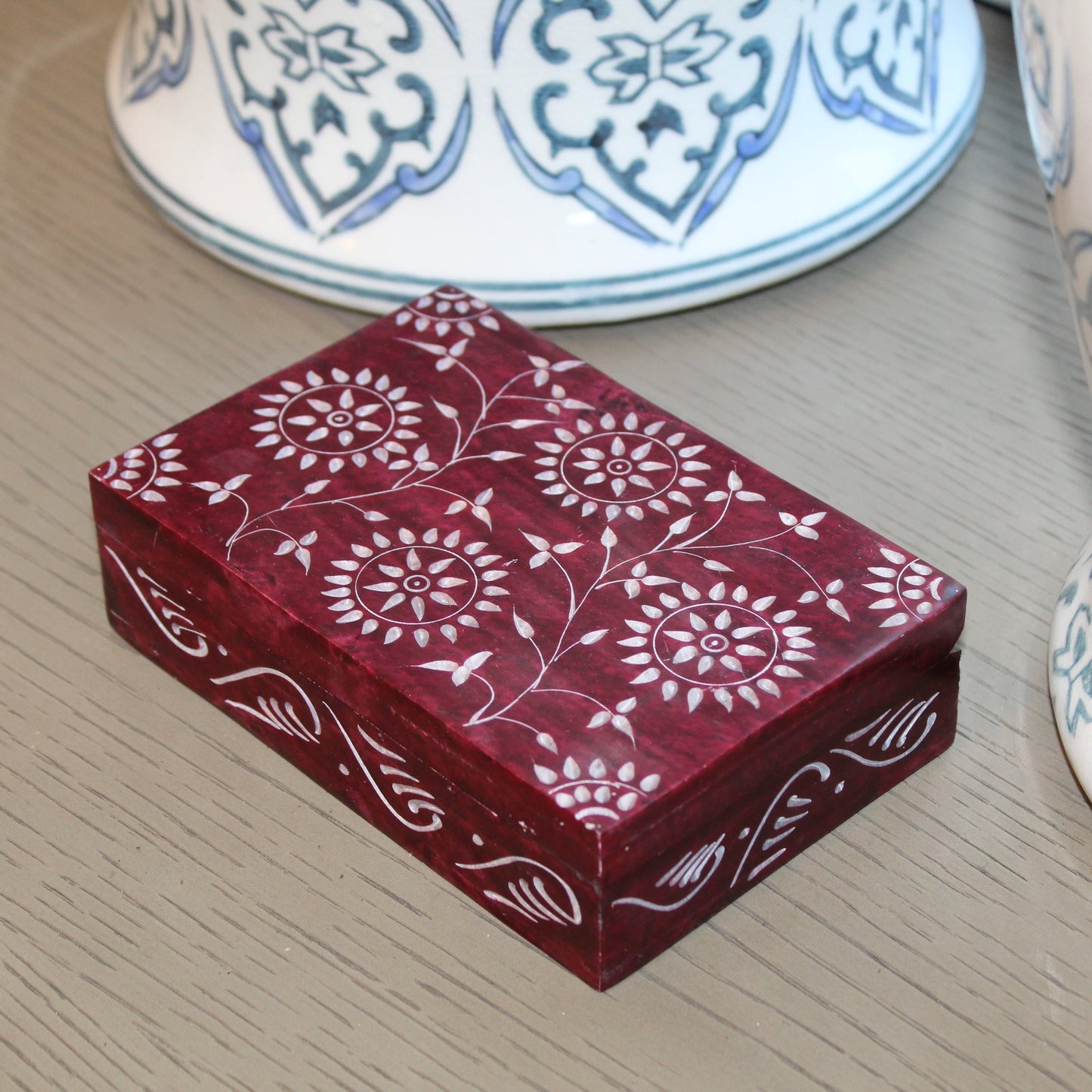 Red decorative box with white floral patterns on a wooden surface with ceramic items in the background.