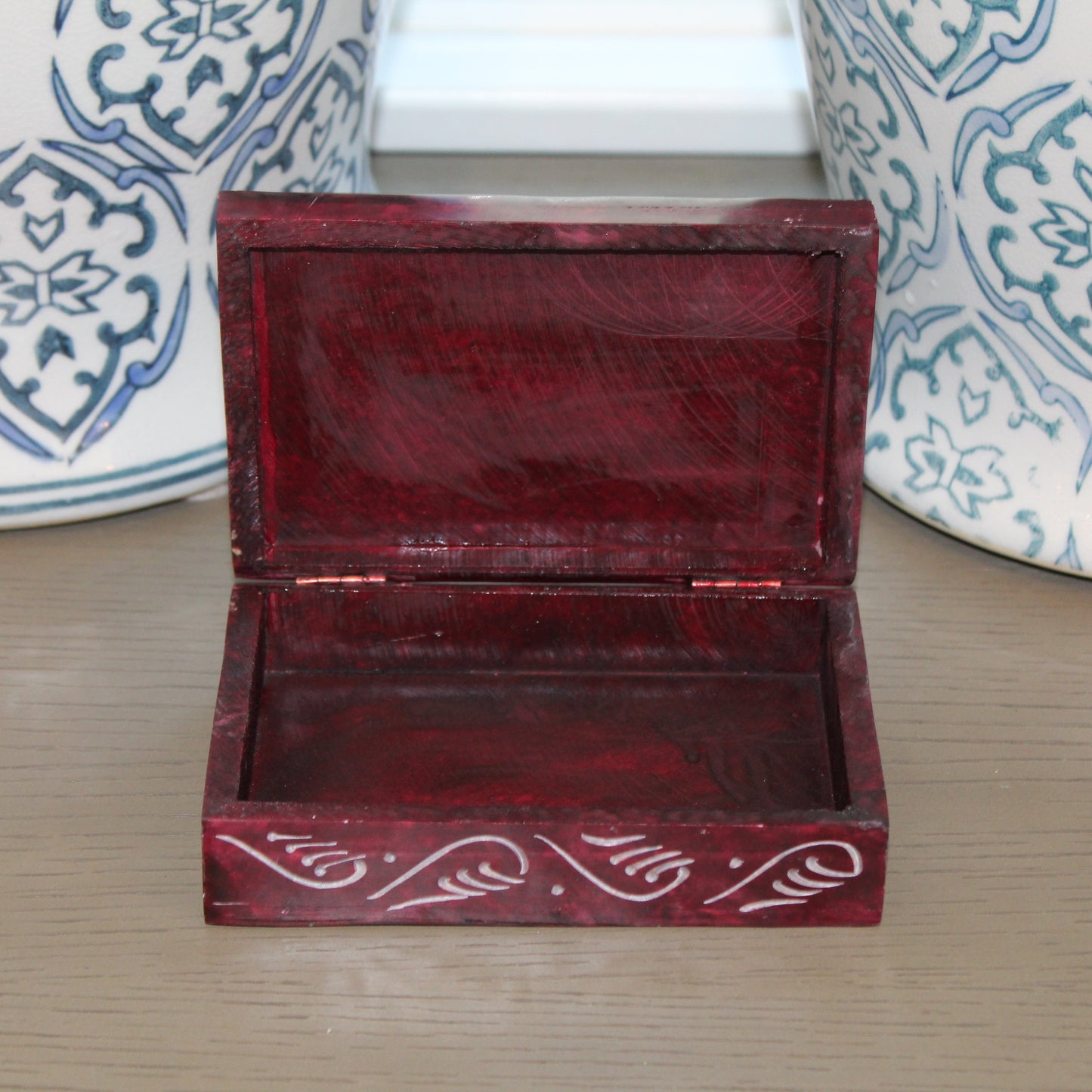 Open red jewelry box with intricate designs on a wooden surface with decorative vases in the background.