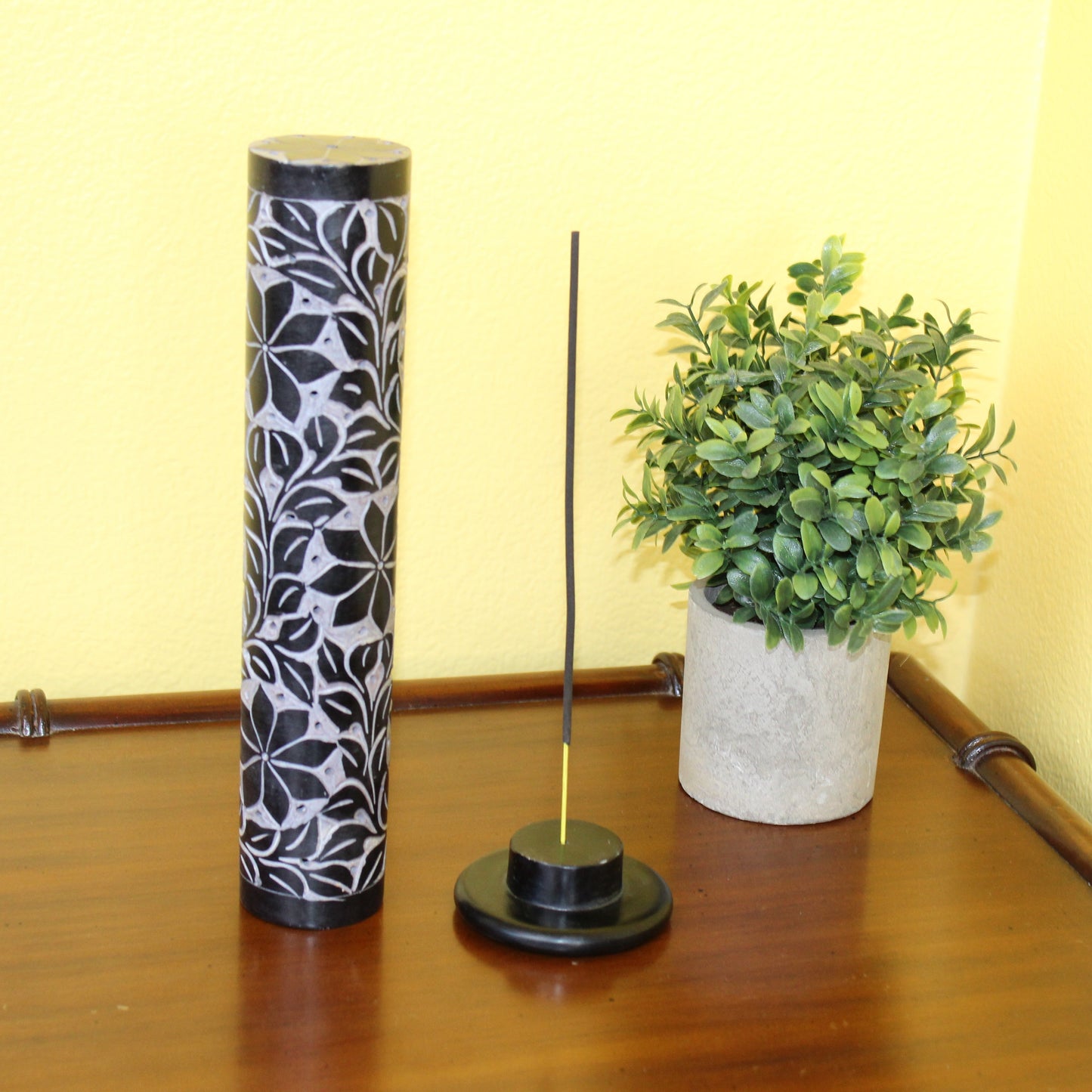 Tall cylindrical container with floral design on a wooden surface next to a small potted plant.