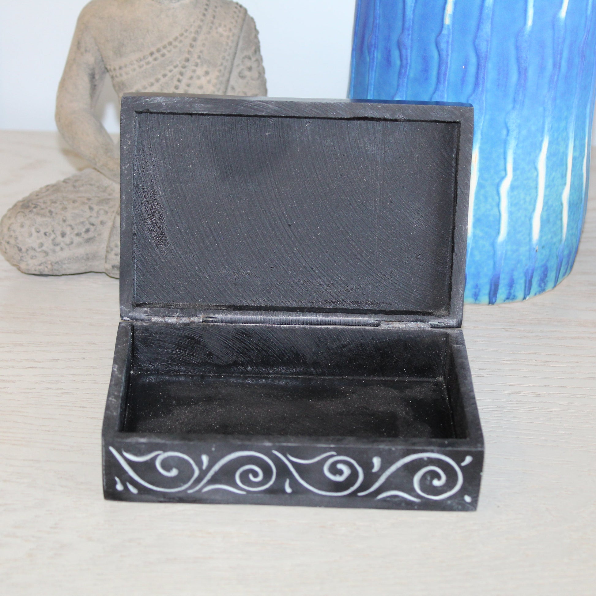 Black decorative box with white patterns on a light surface with a blue vase and stone sculpture in the background.