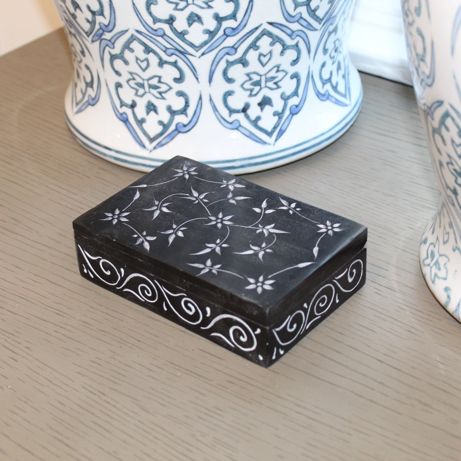 Black decorative box with white patterns on a wooden surface with blue and white ceramic items in the background.