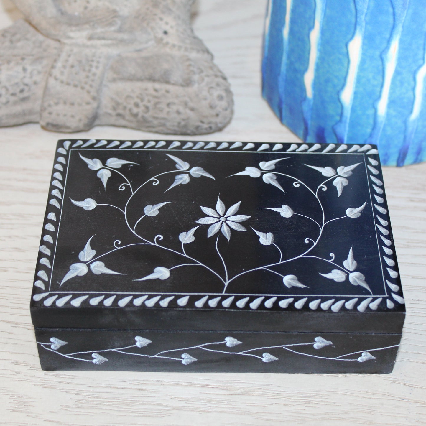 Decorative black box with white floral patterns on a textured surface with a blue candle and stone object in the background.