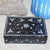 Decorative black box with white floral patterns on a textured surface with a blue candle and stone object in the background.