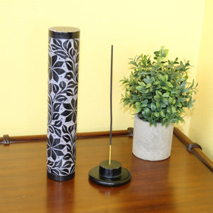 Tall cylindrical container with floral design on a wooden surface next to a small potted plant.